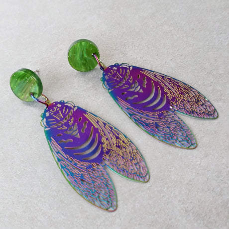 Purple and blue toned cicada-style stainless steel earrings with green acrylic toppers on a light gray background