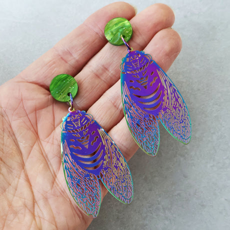 A pair of dangle earrings with stainless steel circada dangles hanging from green sparkle swirl acrylic toppers, being held in a hand. The circadas have purple/blue tones. 