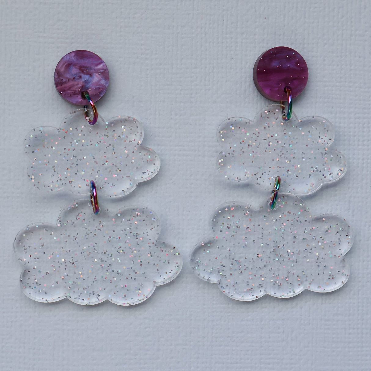 Pair of clear glittery cloud-shaped earrings with purple circular tops on a light gray background.