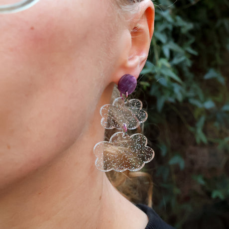 Close-up of a person wearing clear glittery acrylic cloud themed earrings with a purple acrylic topper.
