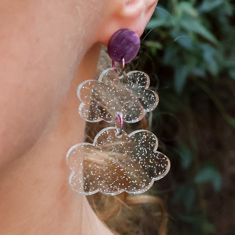 Glittery acrylic cloud dangle earrings hanging from a purple acrylic topper, being modelled