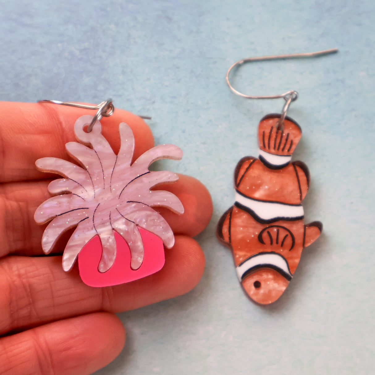 An asymmetrical pair of acrylic earrings with an orange clownfish on one side and a pink sea anemone on the other. Topped with stainless steel french hooks. Being held in a hand