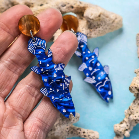 Blue fish-shaped coelocanth earrings with white accents and brown toppers held by a hand against a coral background.