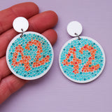 Pair of earrings with colour blindness test design featuring dots in orange and blue showing the number 42, and being held up by a hand on a purple background