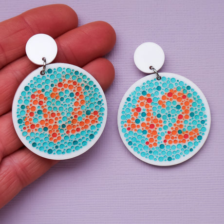Pair of earrings with colour blindness test design featuring dots in orange and blue showing the number 42, and being held up by a hand on a purple background
