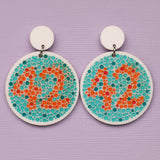 Pair of earrings with colour blindness test design featuring dots in orange and blue showing the number 42 on a purple background