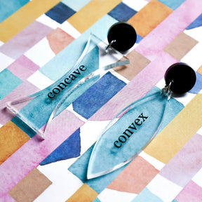  A pair of asymmetrical lens shaped earrings labeled 'concave' on one side and 'convex' on the other on a colorful geometric patterned surface.