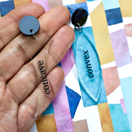 Laser cut acrylic earrings labeled 'concave' and 'convex' held against a colorful geometric pattern. Dangles are clear acrylic, text is black. Dangles hang from black round toppers. 