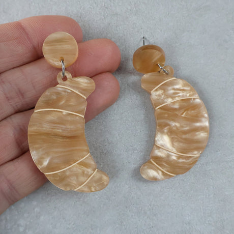 Pair of beige marbled croissant earrings held by a hand on a light grey background