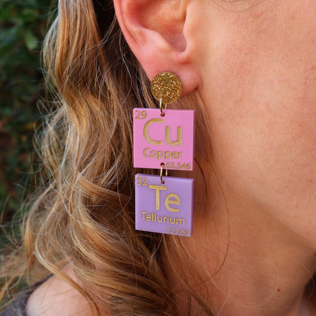 A pair of periodic table earrings spelling CuTe  in purple, pink and gold tones being modelled on an ear