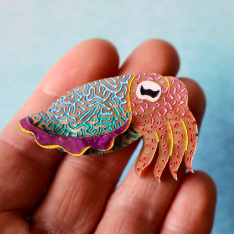 Colourful acrylic cuttlefish brooch held up by a hand against a blue background
