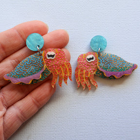 A pair of laser cut acrylic cuttlefish earrings with blue acrylic toppers. Being held in a hand.