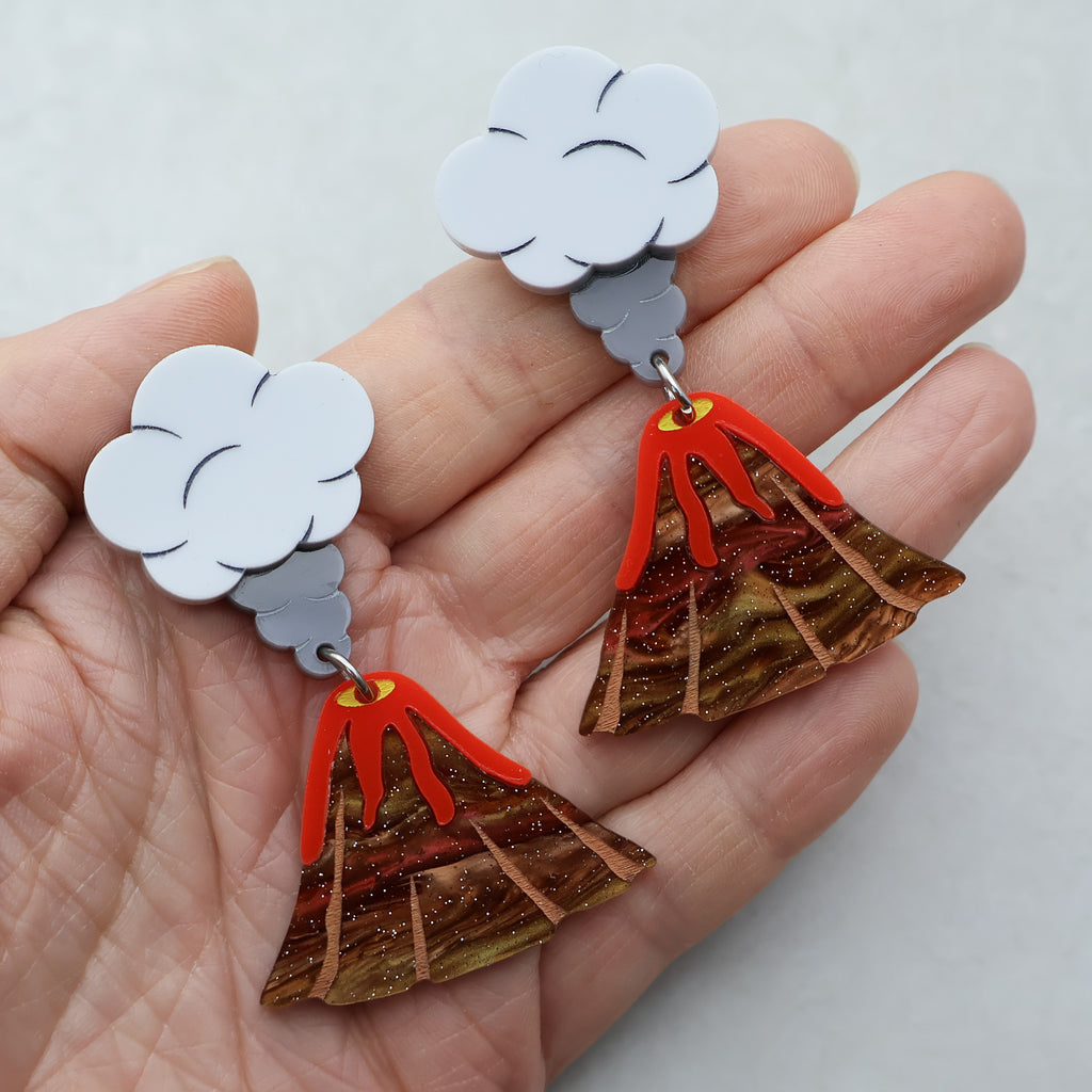 A closeup image of acrylic volcano earrings. The tops of the earrings are grey toned ash clouds, and volcanoes with lava flowing out dangle below. 