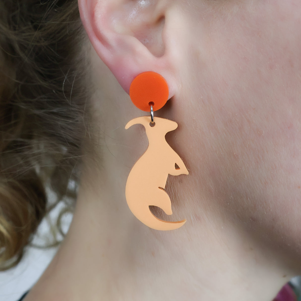 Orange parasaurolophus dangle earrings with darker orange toppers. Made from laser cut acrylic. Being modelled.