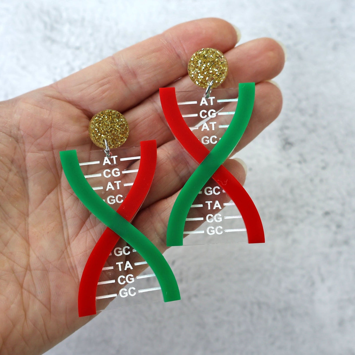 Acrylic DNA strand earrings in Christmas colours, held in a hand against a textured background.