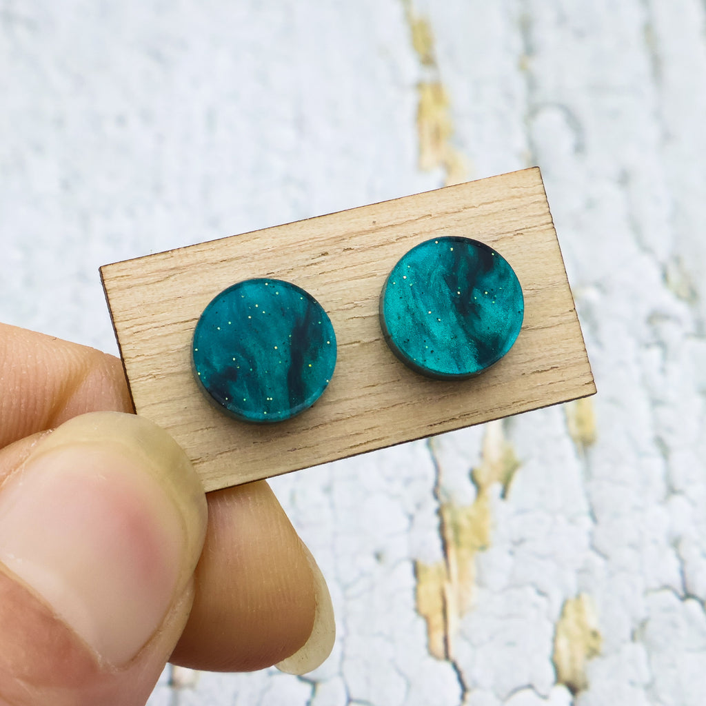 A pair of small swirly green acrylic studs on a wooden backing card.