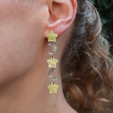 Closeup of a person wearing acrylic star and moon earrings. The stars are yellow and the moons are grey/brown.