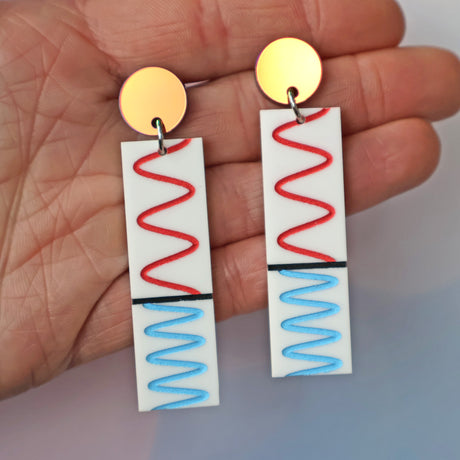 Pair of earrings with red and blue wave designs depicting the doppler effect, held between fingers.