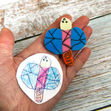 An acrylic dragonfly brooch in the style of a small child's drawing, next to the child's drawing that inspired the design. 