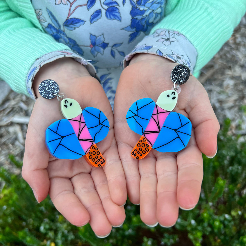 Acrylic dragonfly earrings in the style of a small child's drawing, being held in a small child's hands. Laser cut from acrylic.