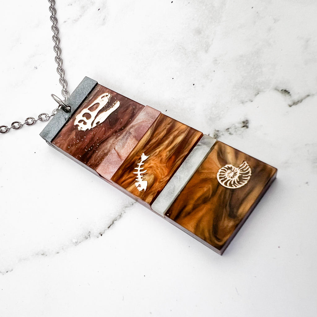 A pendant representing sedimentary layers, rectangular in shape and comprising 6 strips of different grey and brown acrylics, with a t-rex skull, fish bones and nautilus engraved and handpainted in the larger layers. The pendant hangs on a fine stainless steel chain.