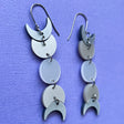 A pair of moon phase earrings displayed against a piece of purple cardstock. The earrings are made from silver pearlescent acrylic and hang from french hooks. 