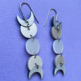 A pair of moon phase earrings displayed against a piece of purple cardstock. The earrings are made from silver pearlescent acrylic and hang from french hooks. 