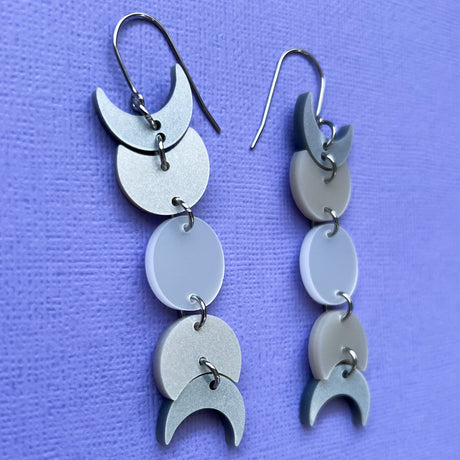 A pair of moon phase earrings displayed against a piece of purple cardstock. The earrings are made from silver pearlescent acrylic and hang from french hooks. 
