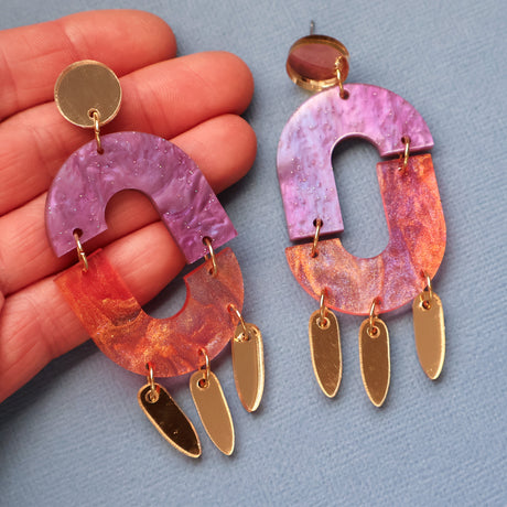 Pair of colourful dangle earrings in pinks and purples with gold accents being held up in a hand on a gray background