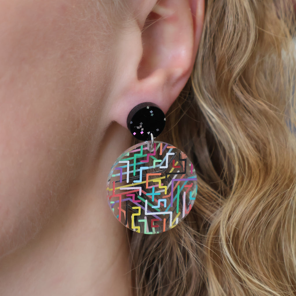 Clear circle earrings with multicoloured circult designs painted on back and front, and hanging from black glittery earring toppers, being modelled.