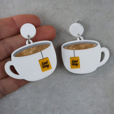 Teacup earrings with 'Earl Grey' tags held by a hand on a gray background