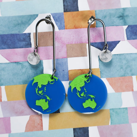Earrings with earth and moon acrylic  dangles hanging on stainless steel arches against a colorful geometric background.