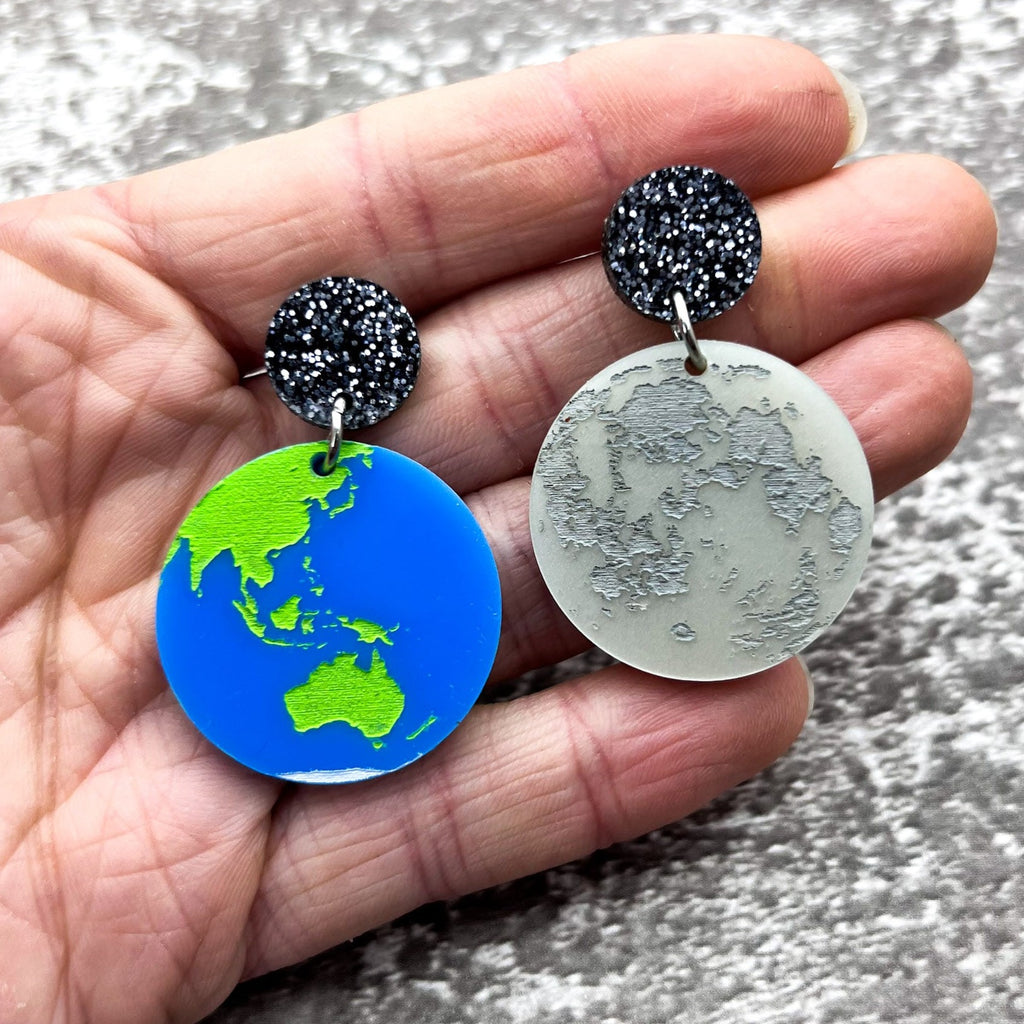 Asymmetrical acrylic earth and moon earrings, hanging from black glitter acrylic toppers.