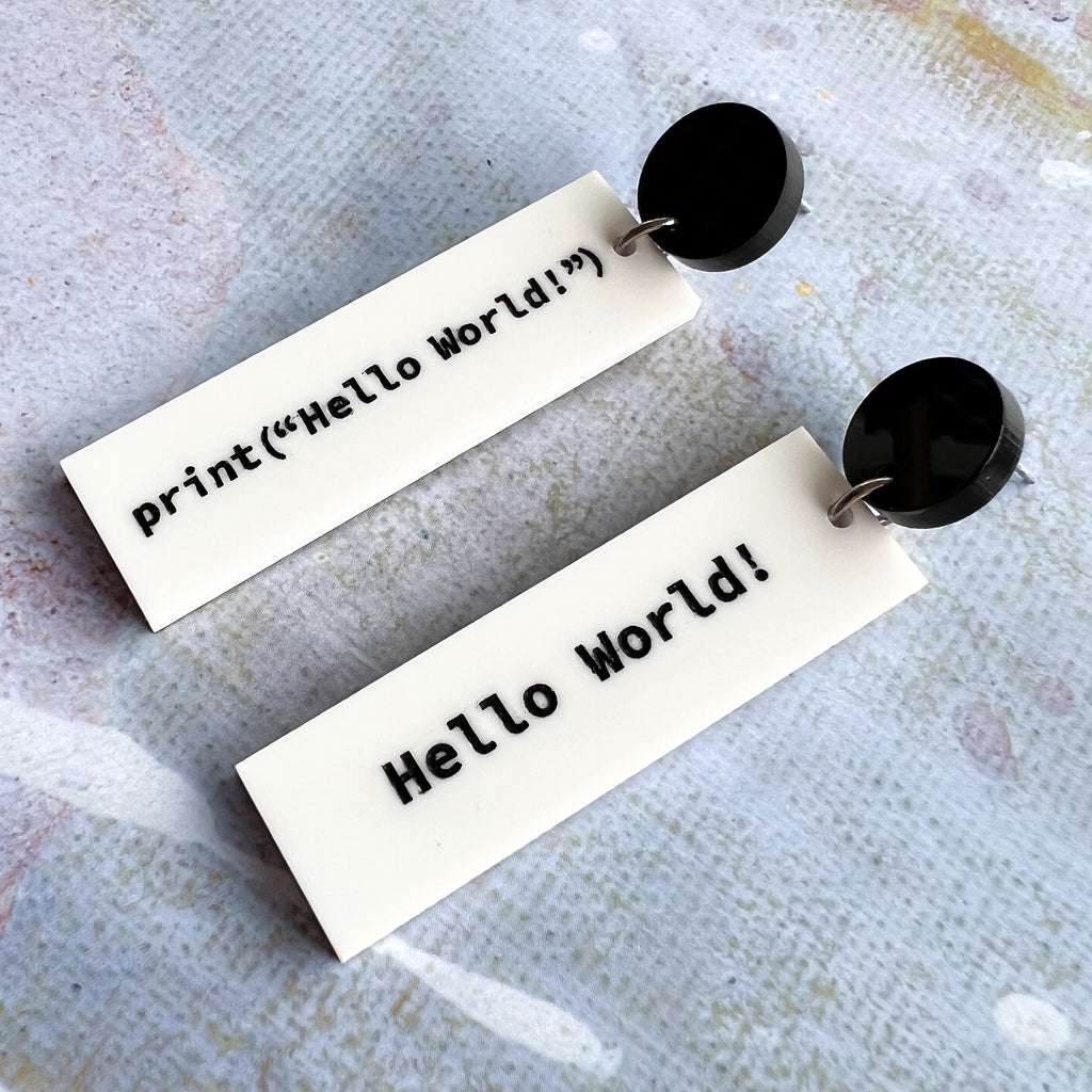 Rectangular white dangle earrings with print("Hello World!")  in black text on one side and 'Hello World!' on the other side. They hang from black acrylic toppers. 