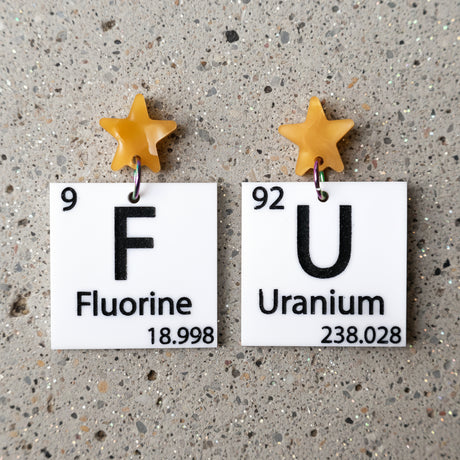 black and white asymmetrical periodic table earrings with 'Fluorine' and 'Uranium' labels, and yellow star toppers. 
