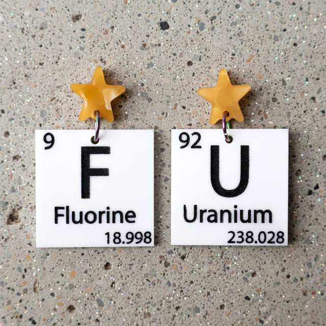 black and white asymmetrical periodic table earrings with 'Fluorine' and 'Uranium' labels, and yellow star toppers. 
