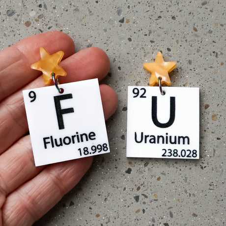 Black and white acrylic earrings with 'Fluorine' and 'Uranium' labels and yellow star toppers. 