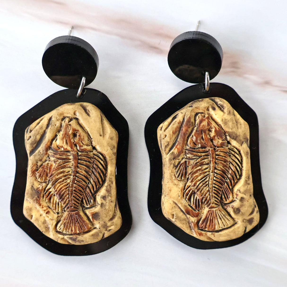 A pair of earrings with a brown fish skeleton cabochon design mounted in a black acrylic border, with black acrylic toppers.