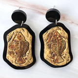 A pair of earrings with a brown fish skeleton cabochon design mounted in a black acrylic border, with black acrylic toppers.