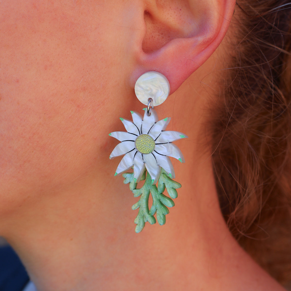 A close-up of a person's ear wearing a dangling earring shaped like a white flower with pale green leaves and a light green center, attached to a round white stud.