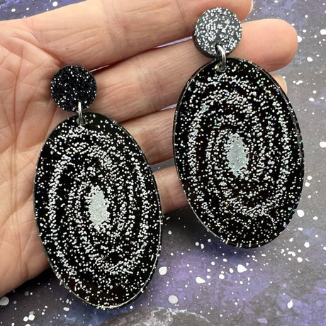 Statement sized milky way galaxy acylic earrings. The earrings are oval in shape and contain silver toned stars handpainted against a black background. The earrings hang from black glitter acrylic toppers. Earrings are being held up for a better view. 