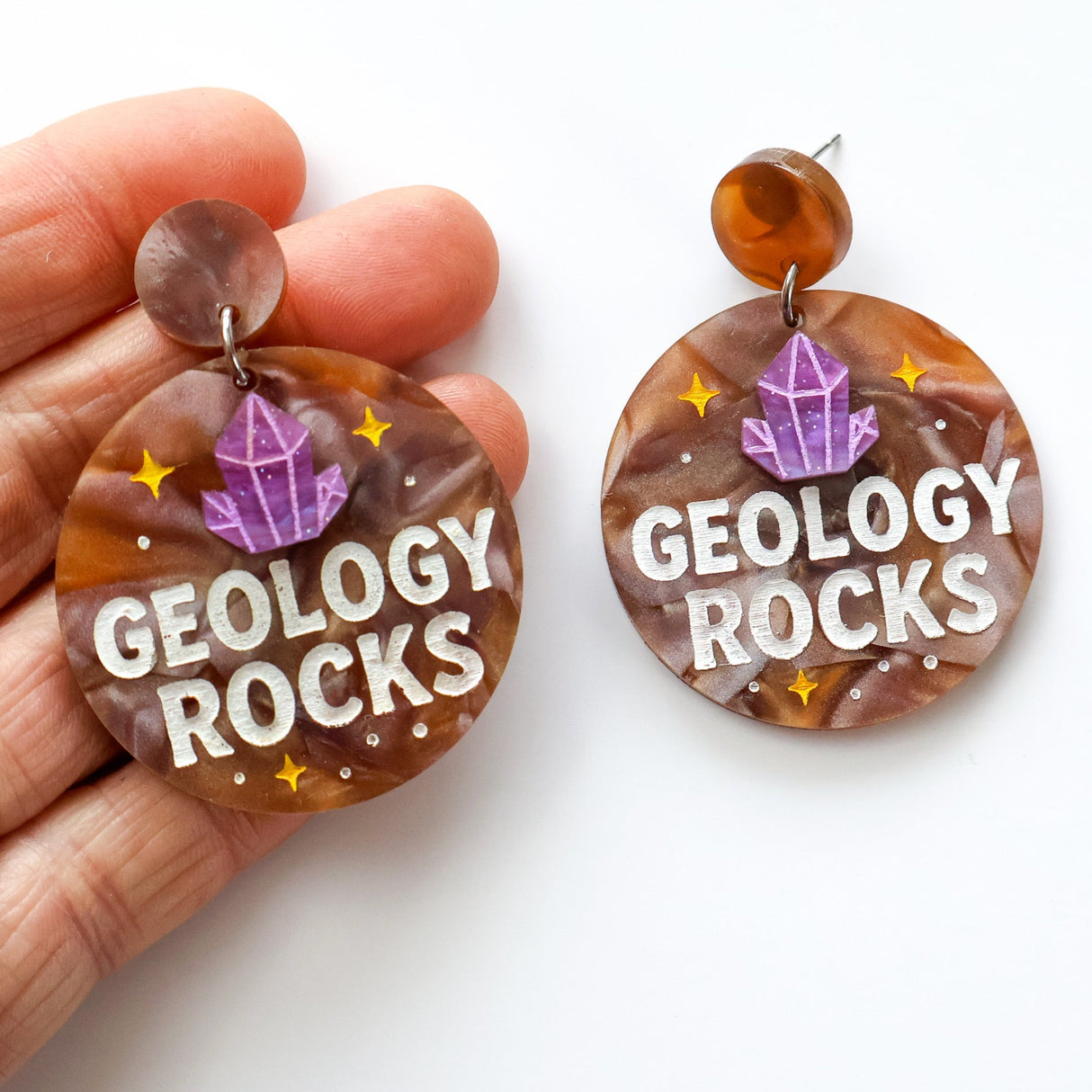 Earrings with 'GEOLOGY ROCKS' text and gemstone design held by a hand on a white background