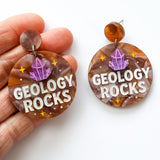 Earrings with 'GEOLOGY ROCKS' text and gemstone design held by a hand on a white background