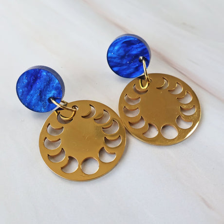 Gold toned round dangle earrings a moon phase design with blue circular acrylic toppers on a white background.