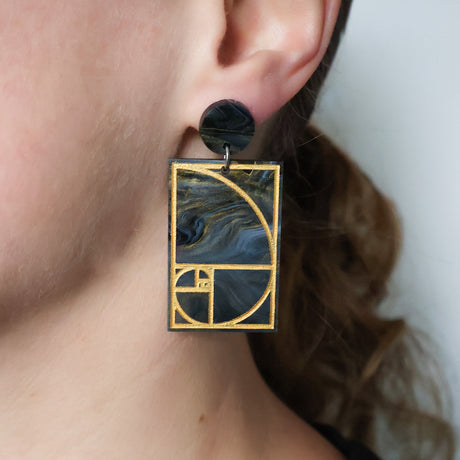 A pair of rectangular acrylic earrings displaying a gold painted golden ratio design in dark grey and gold acrylic, with a matching round acrylic topper, being modelled in an ear. 