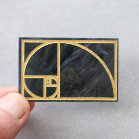 Gold Fibonacci spiral on a black marble textured arylic rectangular brooch. Brooch is being held up in a hand