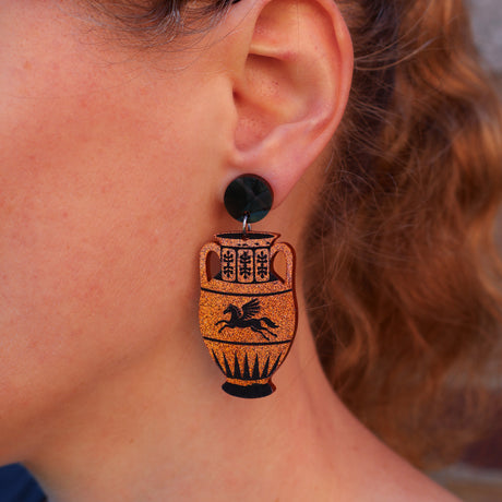 Close-up of a person's ear wearing a dangling earring shaped like an ancient Greek-style amphora jar with black details and a black stud.
