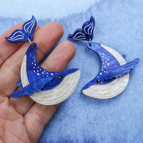Laser cut acrylic humpback whale earrings being held in hand against a blue gradient background.