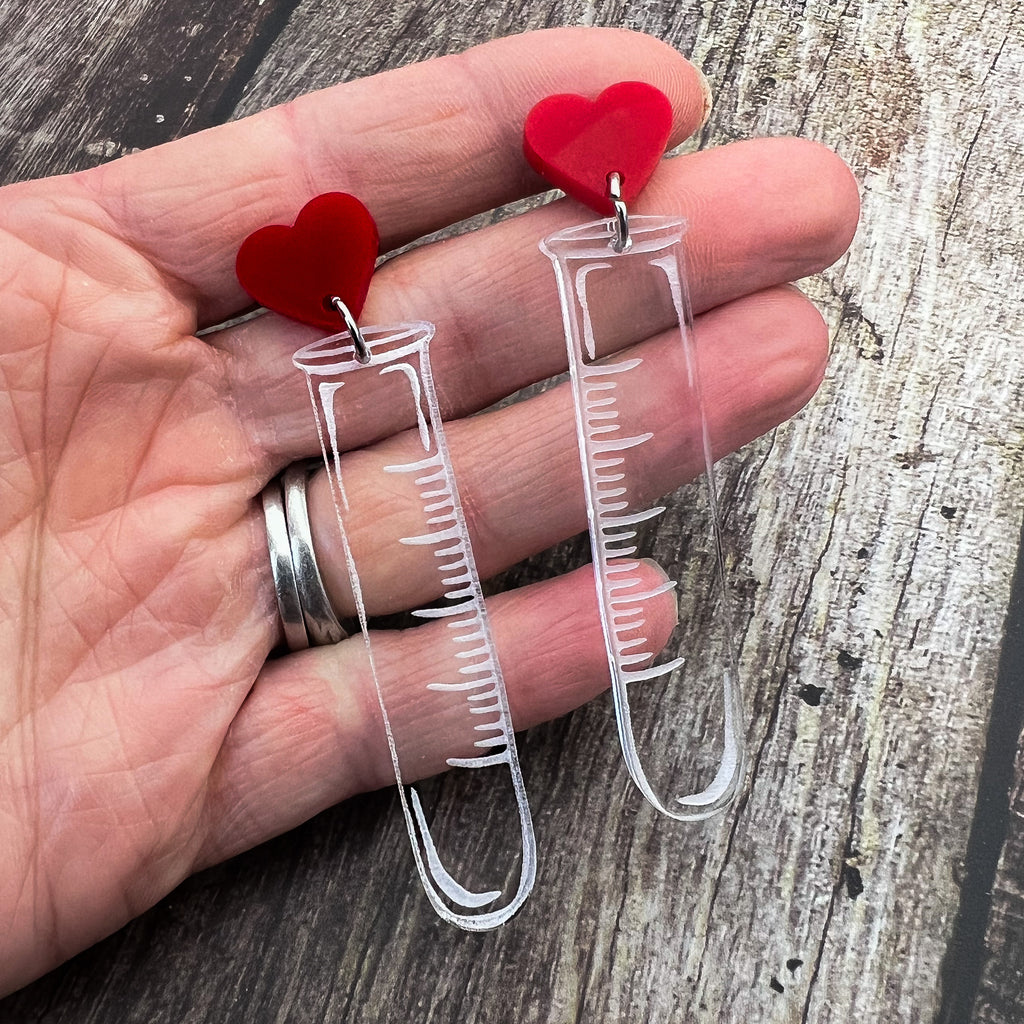 Test tube earrings, laser cut and engraved in clear acrylic, and hanging from red heart earring toppers. 