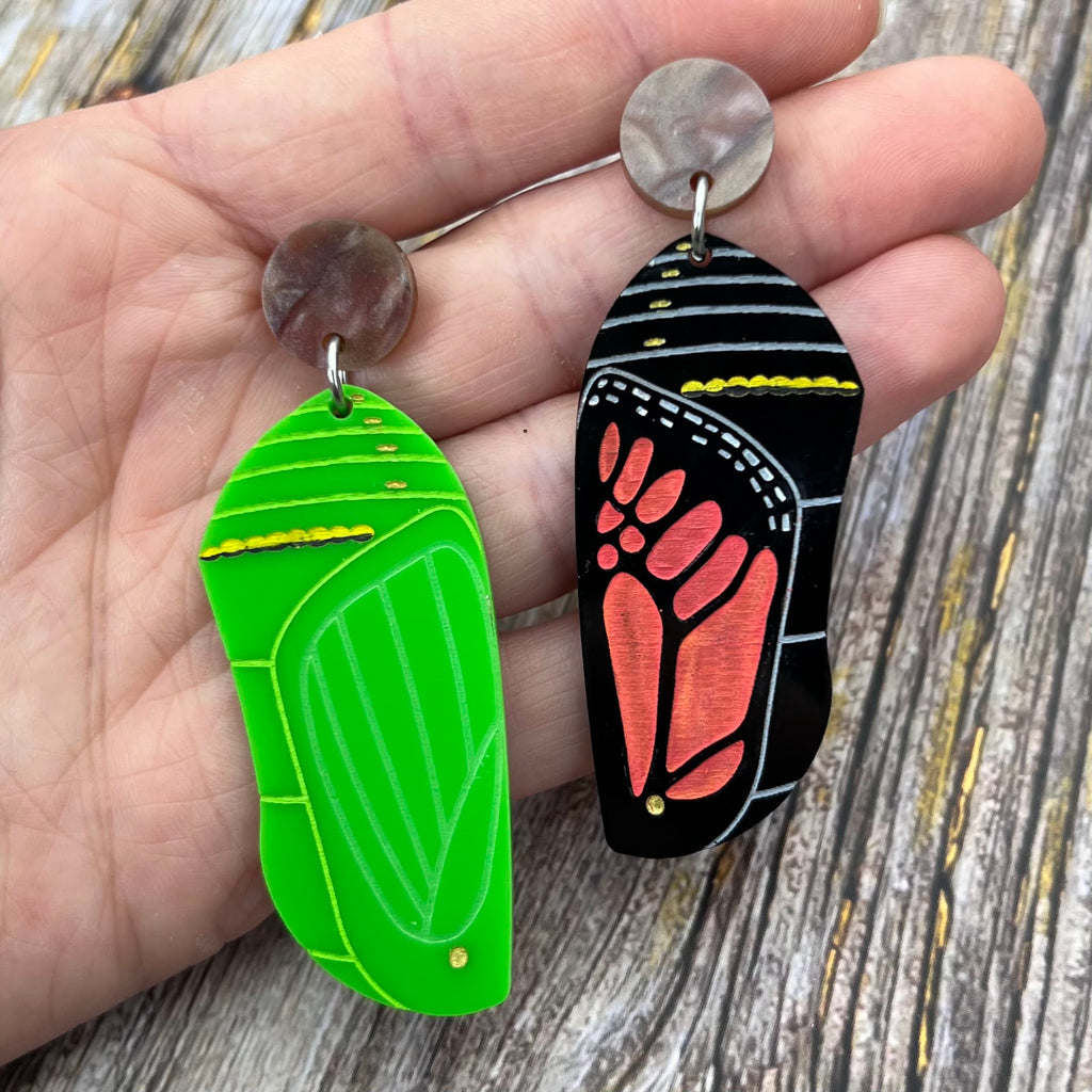 Asymmetrical earrings of a monarch butterfly chrysalis, with one side being a green chrysalis with the other being a late stage black and red chrysalis, showing the different stages of development. 